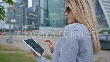 一个年轻的金发女孩在市中心摩天大楼的背景上使用平板电脑。